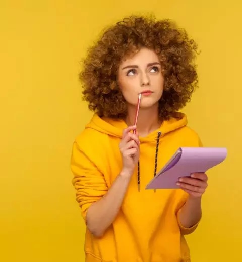Młoda dziewczyna z burzą loków na głowie, ubraną w żółtą bluzę, stoi na żółtym tle. W lewej ręce trzyma notatnik, w prawej ołówek. Oczy ma skierowane ku górze, zastanawia się nad czymś i robi z tego notatki.