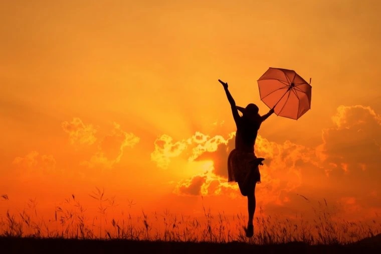 Pomarańczowo czarne zdjęcie dziewczyny z otwartą parasolką trzymaną w dłoni, na tle zachodzącego słońca. Rozłożone ręce postaci oraz ciało w podskoku wskazuje na bardzo radosny nastrój.