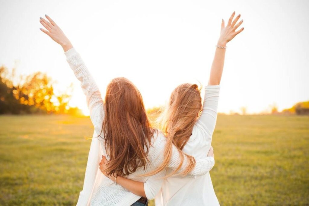 Dwie młode kobiety stojące na polanie tyłem do aparatu, na tle zachodzącego słońca. Dziewczyny obejmują się jedną ręką w pasie a druga mają wyprostowaną i uniesioną nad głowy do nieba.